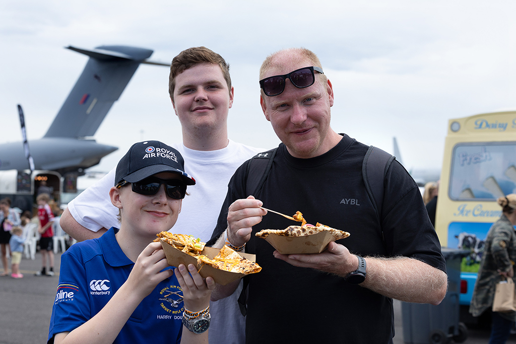 Three smiling males of different ages posing together while eating fastfood from paper trays at the event, with a Royal Air Force Atlas aircraft visible in the background.